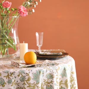Puede incluir: Una mesa de comedor con un mantel floral blanco, con estampados florales verdes y azules. La mesa est&aacute; puesta con platos, servilletas, una vela, un jarr&oacute;n de cristal con flores rosas y una naranja.