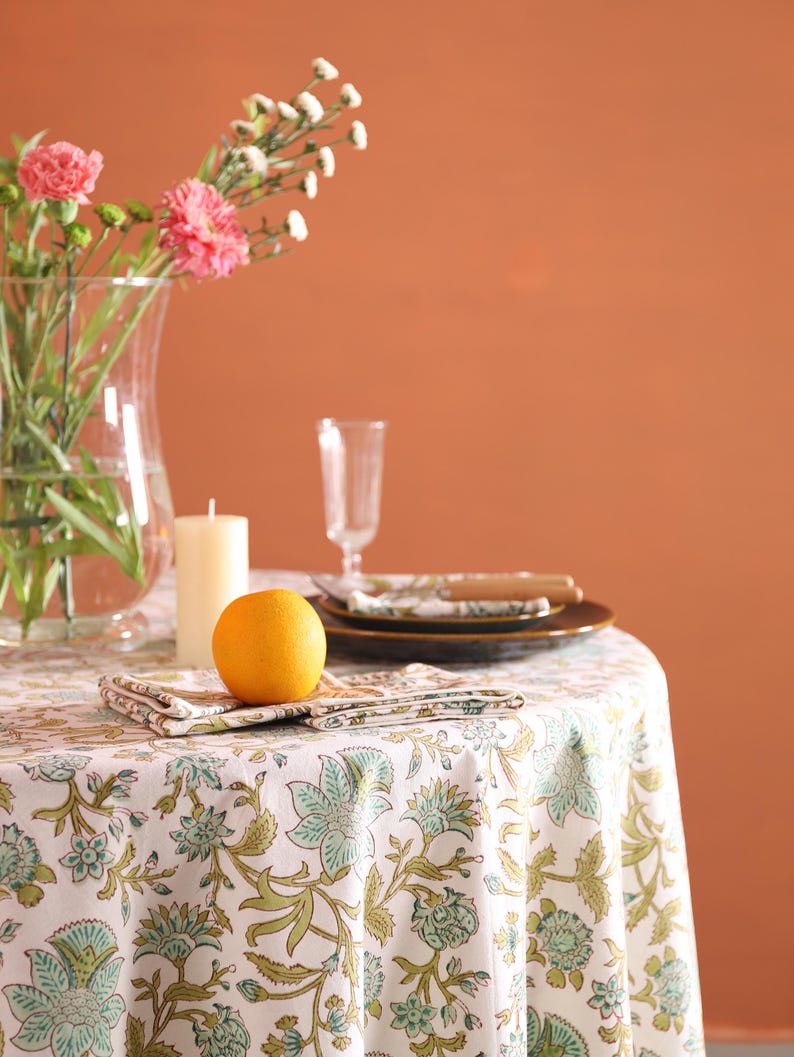 Puede incluir: Una mesa de comedor con un mantel floral blanco, con estampados florales verdes y azules. La mesa est&aacute; puesta con platos, servilletas, una vela, un jarr&oacute;n de cristal con flores rosas y una naranja.