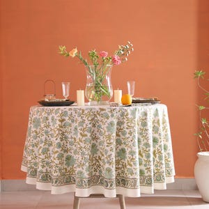 Puede incluir: Una mesa redonda preparada para cenar, cubierta con un mantel blanco con un estampado floral verde y dorado. La mesa est&aacute; puesta con platos, vasos, velas y un jarr&oacute;n con flores. Tambi&eacute;n hay una fruta naranja en la mesa.