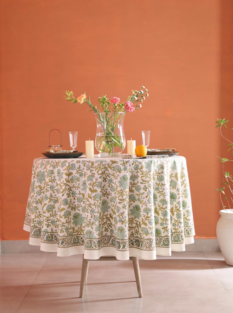 Puede incluir: Una mesa redonda preparada para cenar, cubierta con un mantel blanco con un estampado floral verde y dorado. La mesa est&aacute; puesta con platos, vasos, velas y un jarr&oacute;n con flores. Tambi&eacute;n hay una fruta naranja en la mesa.