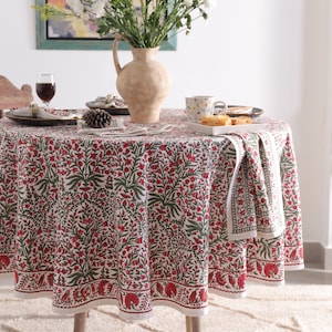 May include: A circular tablecloth featuring a floral design in red and green on a cream backdrop. The table is set with plates, a wine glass, and a vase of flowers. A wooden chair is to the side.