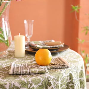 May include: A dining table set with a green and white patterned tablecloth. An orange sits on the table, alongside folded napkins, a candle, and a vase of pink flowers. Brown plates and cutlery are also visible.