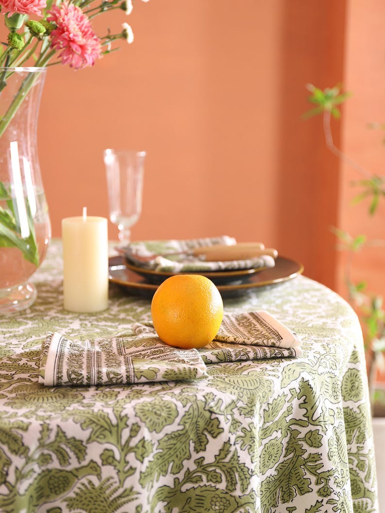 May include: A dining table set with a green and white patterned tablecloth. An orange sits on the table, alongside folded napkins, a candle, and a vase of pink flowers. Brown plates and cutlery are also visible.