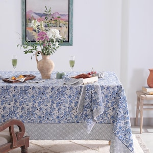 May include: A dining table set with a blue floral tablecloth, matching napkins, and a decorative border. A vase of flowers, a painting, and food items are on the table. The room has a rug and a wooden chair.