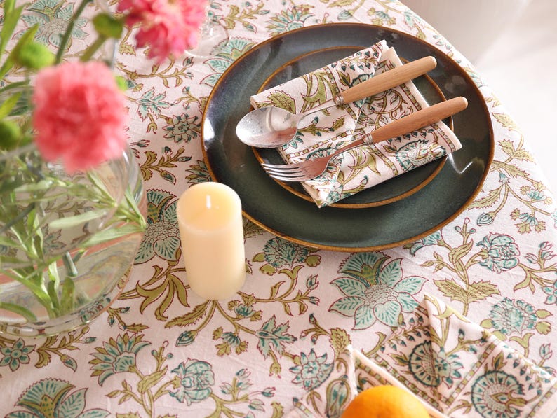 Puede incluir: Una mesa puesta con un plato verde oscuro, una servilleta doblada con un dise&ntilde;o floral verde y cubiertos con mangos de madera. Una vela encendida y un jarr&oacute;n con flores rosas se encuentran sobre un mantel floral.