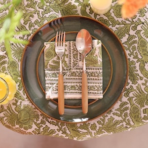 May include: A dining table setting featuring a green plate, a fork, and a spoon with wooden handles. A patterned napkin sits on the plate. The table is covered with a green and white floral tablecloth. A candle and a glass are also visible.