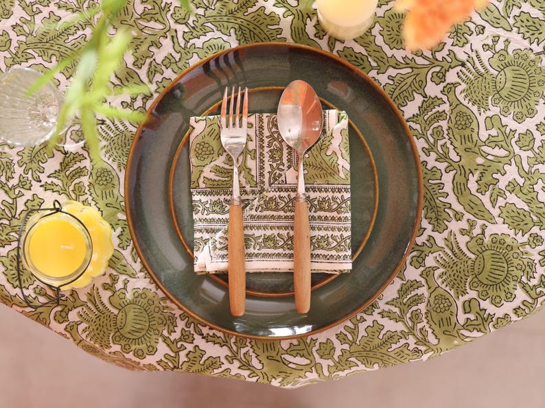 May include: A dining table setting featuring a green plate, a fork, and a spoon with wooden handles. A patterned napkin sits on the plate. The table is covered with a green and white floral tablecloth. A candle and a glass are also visible.