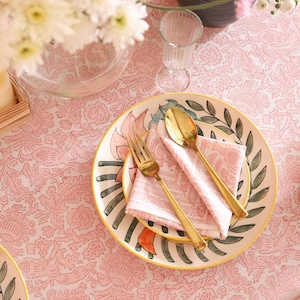 May include: A table setting with a pink floral patterned tablecloth. A plate with a gold fork and spoon, and a pink napkin. A vase of white flowers, candles, and a pomegranate are also on the table.