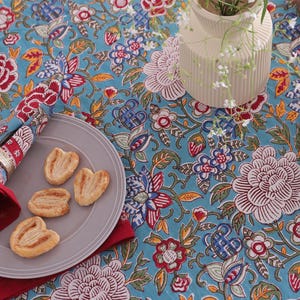 May include: A table setting with a teal floral tablecloth, a gray plate with pastries, and a red and blue napkin. A gold fork and a white vase with flowers are also visible.