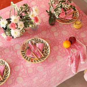 May include: Pink tablecloth with a floral pattern and white accents. The tablecloth is set with plates, napkins, and cutlery. There are also flowers in a vase on the table.