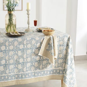 May include: A light blue tablecloth with a floral pattern and a beige border. A matching napkin is folded on the table. The table is set with plates, a candle holder, a vase of flowers, and a glass of red liquid.