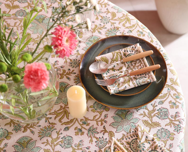 Puede incluir: Una mesa puesta con un mantel floral, un plato verde oscuro y una servilleta estampada. Cubiertos con mangos de madera descansan sobre la servilleta. Un jarr&oacute;n de cristal contiene flores rosas y una vela color crema.