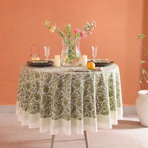 May include: A round dining table with a green floral tablecloth. The table is set with plates, glasses, candles, and a vase of flowers. An orange is placed on the table. The table has light-colored wooden legs.
