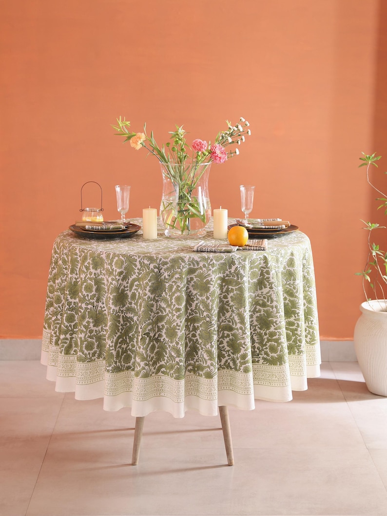 May include: A round dining table with a green floral tablecloth. The table is set with plates, glasses, candles, and a vase of flowers. An orange is placed on the table. The table has light-colored wooden legs.