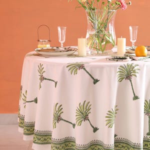 May include: A white round tablecloth with a green palm tree pattern and a green border. The table is set with plates, glasses, candles, and a vase of flowers. An orange sits on the table.