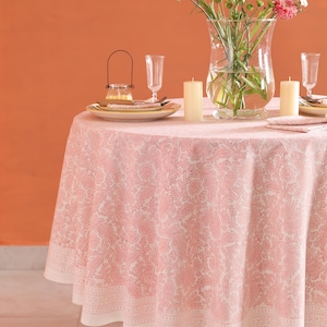 May include: A round tablecloth with a pink floral pattern and a white border is set for a meal. The table is decorated with a vase of flowers, candles, plates, and glasses. The background is a solid orange color.