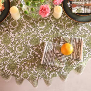 May include: A round tablecloth with a green and white floral design, set for a meal. Includes green plates, napkins, candles, and an orange. The table setting has a rustic, natural aesthetic.