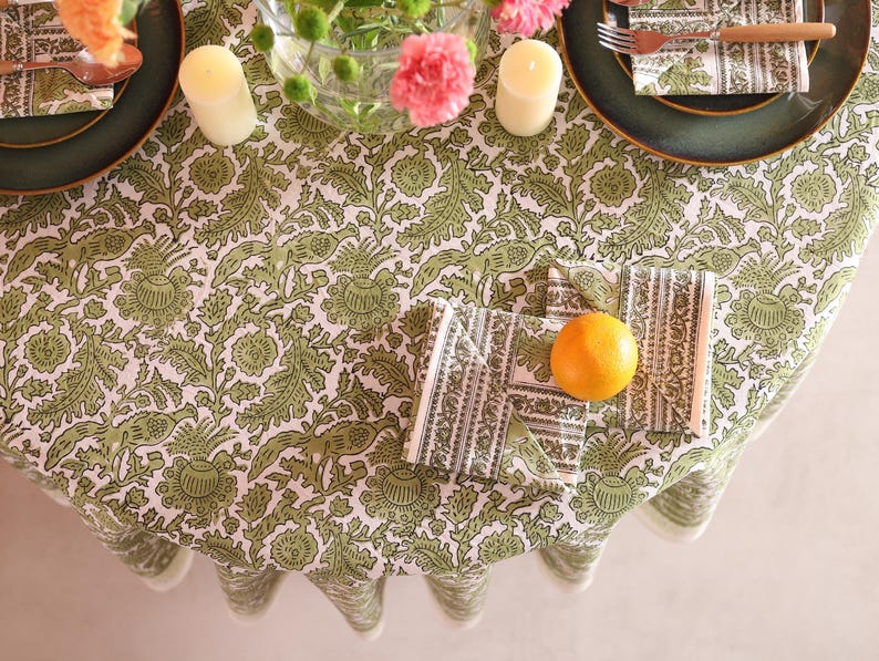 May include: A round tablecloth with a green and white floral design, set for a meal. Includes green plates, napkins, candles, and an orange. The table setting has a rustic, natural aesthetic.