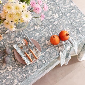 May include: A table setting with a blue and white paisley tablecloth, featuring a plate, silverware, and a patterned napkin. Two pomegranates and a vase of flowers are also on the table. Two candles are in a wooden tray.