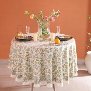 Puede incluir: Una mesa redonda para dos, con un mantel estampado floral en blanco, verde y dorado. La mesa est&aacute; puesta con platos, vasos, velas y un jarr&oacute;n con flores. Una naranja est&aacute; sobre la mesa.
