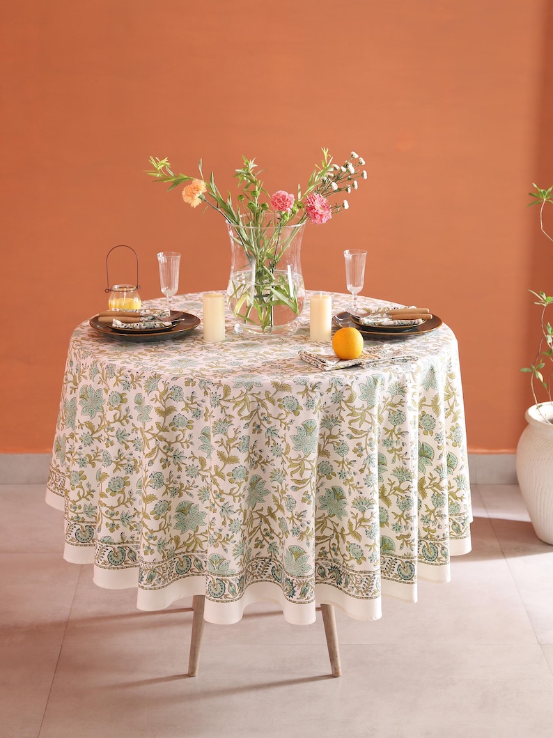 Puede incluir: Una mesa redonda para dos, con un mantel estampado floral en blanco, verde y dorado. La mesa est&aacute; puesta con platos, vasos, velas y un jarr&oacute;n con flores. Una naranja est&aacute; sobre la mesa.