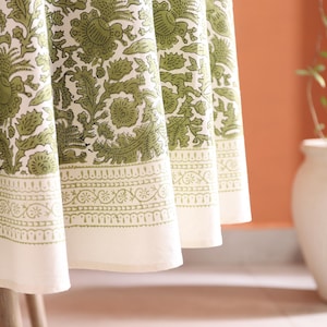 May include: Close-up of a white tablecloth with a green floral pattern. The tablecloth has a decorative border with a repeating design. The fabric is draped over a table, with the edge of the tablecloth visible.