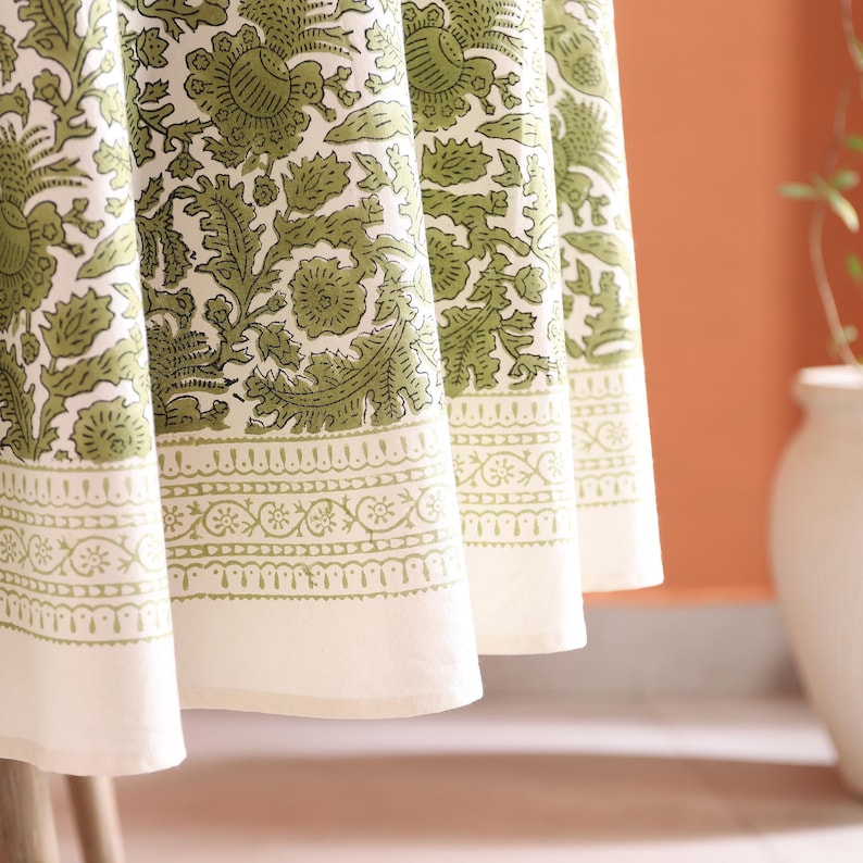 May include: Close-up of a white tablecloth with a green floral pattern. The tablecloth has a decorative border with a repeating design. The fabric is draped over a table, with the edge of the tablecloth visible.