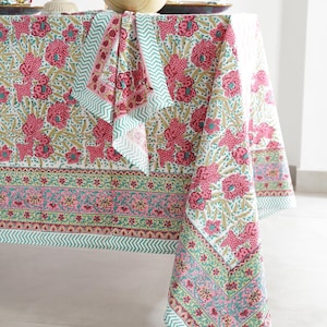 Puede incluir: Un mantel blanco con un vibrante estampado floral en rosa, verde y dorado. El mantel presenta un diseño repetido de grandes flores y hojas, con un borde contrastante y una servilleta a juego. La tela está drapeada sobre una mesa.