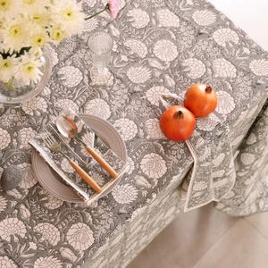 May include: A gray floral tablecloth with white flower patterns covers a table. A place setting with a plate, silverware, and a napkin is visible. Two pomegranates and a vase of flowers are also on the table.