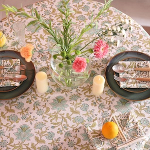 Puede incluir: Una mesa puesta para dos con un mantel floral, platos verdes y cubiertos con mango de madera. Tambi&eacute;n hay servilletas florales, un jarr&oacute;n con flores, dos velas y un peque&ntilde;o candelabro. Una naranja descansa sobre una servilleta.