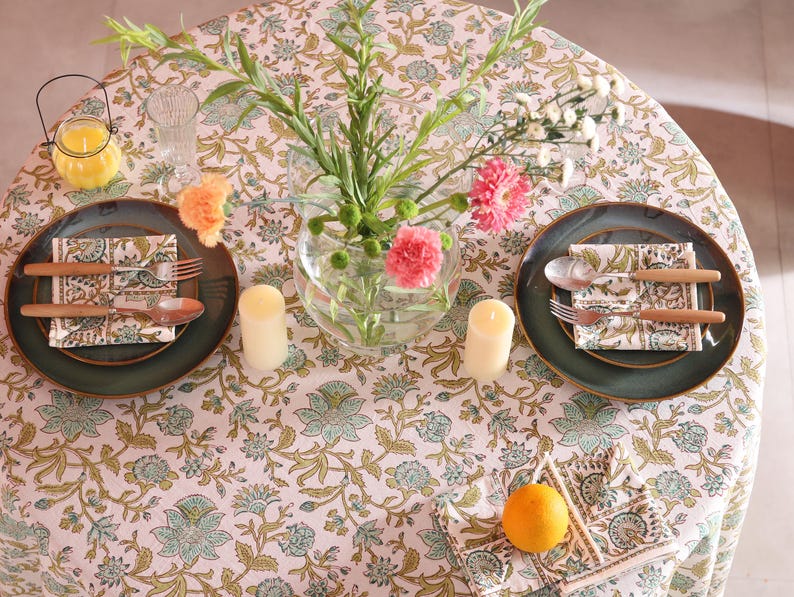 Puede incluir: Una mesa puesta para dos con un mantel floral, platos verdes y cubiertos con mango de madera. Tambi&eacute;n hay servilletas florales, un jarr&oacute;n con flores, dos velas y un peque&ntilde;o candelabro. Una naranja descansa sobre una servilleta.