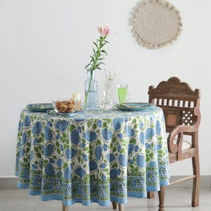 May include: A round tablecloth with a blue floral pattern, set for a meal. The table is set with plates, a glass bowl of snacks, and a vase with a pink flower. A wooden chair is next to the table.