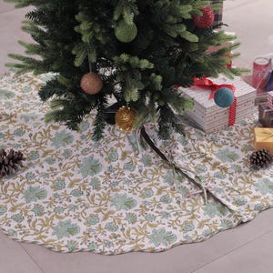 May include: A Christmas tree skirt with a floral pattern in shades of green and tan. The skirt is beneath a decorated Christmas tree with ornaments and wrapped gifts. A pine cone and candles are also visible.