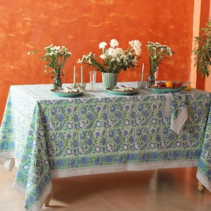 Op de afbeelding: Een wit tafelkleed met een groen en blauw bloemenpatroon. Het tafelkleed ligt op een tafel met een vaas met witte bloemen en een kom met fruit.