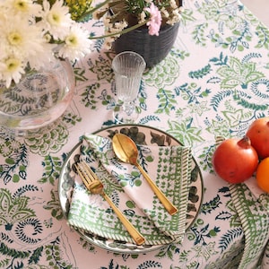 May include: A table setting with a white and green floral patterned tablecloth. A plate with a gold fork and spoon, a napkin, and a glass are set. Fresh flowers and fruit, including pomegranates and an orange, are also present.