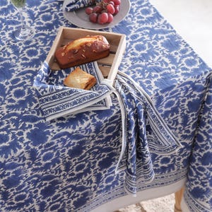 Può includere: Un tavolo è apparecchiato con una tovaglia a motivi floreali blu e bianchi. Un vassoio di legno contiene pane e un pasticcino, con un tovagliolo abbinato. Anche l'uva e un bicchiere sono sul tavolo.