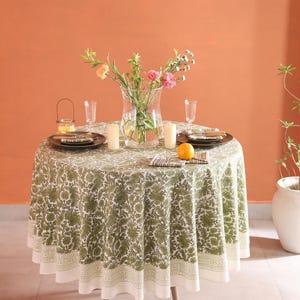 May include: A round dining table with a green floral tablecloth, set with plates, glasses, and candles. A vase of flowers is in the centre, with an orange on the table. The table is supported by three wooden legs. The tablecloth has a white border.