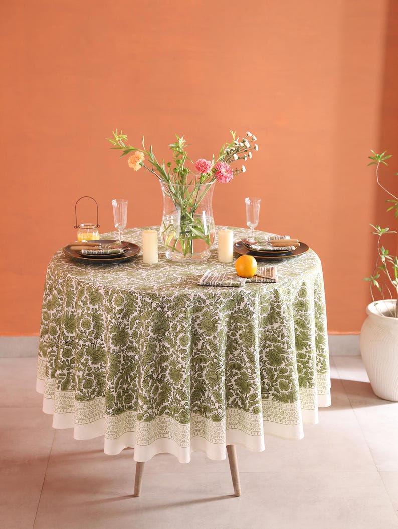 May include: A round dining table with a green floral tablecloth, set with plates, glasses, and candles. A vase of flowers is in the centre, with an orange on the table. The table is supported by three wooden legs. The tablecloth has a white border.