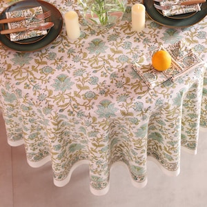 Puede incluir: Un mantel redondo con un estampado floral en tonos verdes y dorados, preparado para una comida. La mesa est&aacute; puesta con platos verdes, cubiertos y servilletas. Una naranja est&aacute; sobre una servilleta. Tambi&eacute;n hay dos velas y un jarr&oacute;n sobre la mesa.