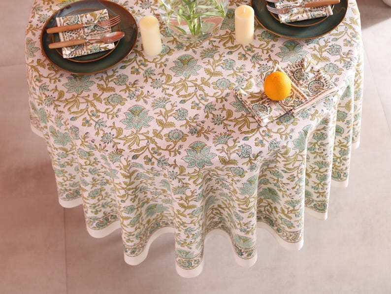 Puede incluir: Un mantel redondo con un estampado floral en tonos verdes y dorados, preparado para una comida. La mesa est&aacute; puesta con platos verdes, cubiertos y servilletas. Una naranja est&aacute; sobre una servilleta. Tambi&eacute;n hay dos velas y un jarr&oacute;n sobre la mesa.