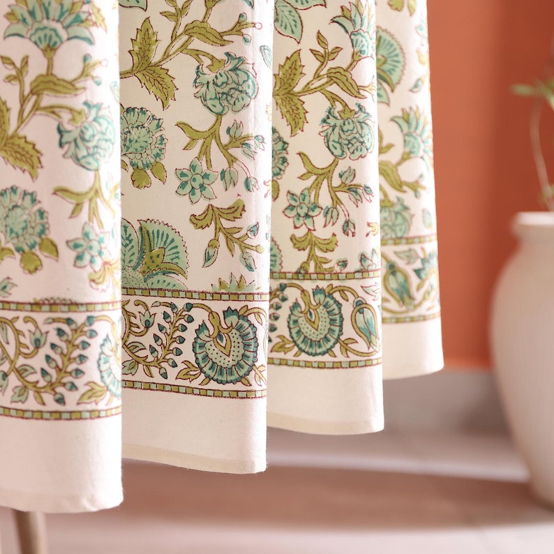 Puede incluir: Primer plano de una tela color crema con un estampado floral repetido en tonos verdes y turquesas. El dise&ntilde;o incluye flores y hojas estilizadas, con un borde detallado en la parte inferior. La tela parece ser una cortina o un mantel.