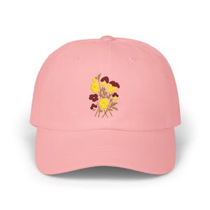 May include: A light pink baseball cap with a floral embroidery design. The embroidered flowers are yellow, maroon, and light brown. The cap is made of a soft material and has a curved brim.
