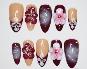Può includere: Un set di dieci unghie finte in varie forme e design. Alcune unghie sono nude con un motivo floreale bianco, mentre altre sono bordeaux con un vortice bianco o un fiore 3D. Accenti dorati sono presenti su diverse unghie.