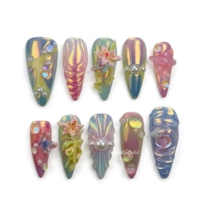 May include: A set of ten iridescent, almond-shaped artificial nails. Each nail features unique designs, including pearls, floral accents, and textured patterns. The color palette includes shades of pink, blue, yellow, and green, creating a shimmering effect.