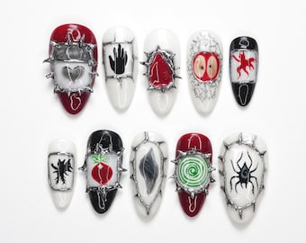 Puede incluir: Un conjunto de diez uñas artificiales con varios diseños. Las uñas presentan una mezcla de colores, incluyendo rojo, blanco y negro, con detalles metálicos plateados. Los diseños incluyen corazones, manos, manzanas y una araña.