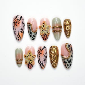 May include: A set of ten artificial nails with various designs. Some nails feature a pink base with black and orange patterns, while others have gold embellishments, floral accents, and a green-to-pink gradient. The nails are oval-shaped.