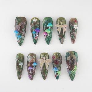 Peut inclure: Dix faux ongles longs et pointus. Chaque ongle présente un design unique avec des éléments sur le thème de la forêt, notamment des champignons, des fleurs et des vignes. La palette de couleurs comprend des verts, des bruns et des touches de bleu, de rose et de blanc.