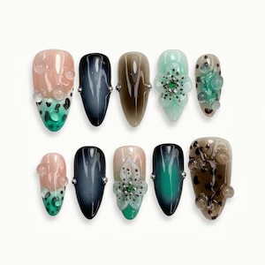 May include: A collection of ten artificial nails in various designs. The nails feature a range of colors, including pink, black, brown, and teal. Some nails have floral or bubble-like embellishments, while others have a gradient effect.