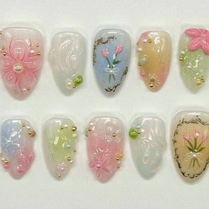 Puede incluir: Un conjunto de diez uñas artificiales con diseños florales. Cada uña presenta un diseño diferente, incluyendo flores en relieve, tulipanes pintados y adornos decorativos en rosa, blanco, azul y verde. También hay cuentas y detalles dorados.