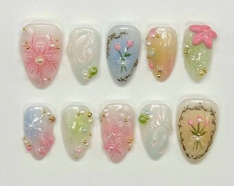 Op de afbeelding: Een set van tien kunstnagels met bloemmotieven. Elke nagel heeft een ander ontwerp, waaronder reliëfbloemen, geschilderde tulpen en decoratieve versieringen in roze, wit, blauw en groen. Goudkleurige kralen en accenten zijn ook aanwezig.
